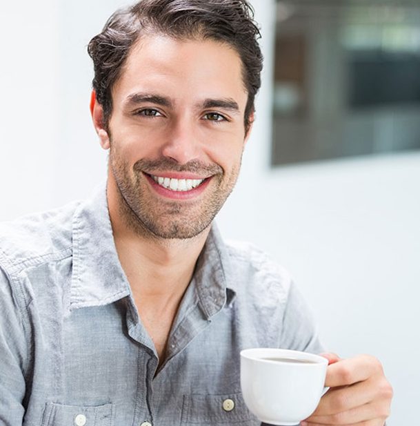Man in light blue shirt smiling while holding white coffee mug indoors