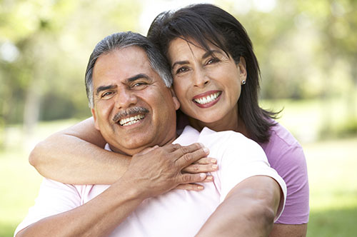 Smiling couple embracing outdoors in casual white and purple clothing against blurred green background.