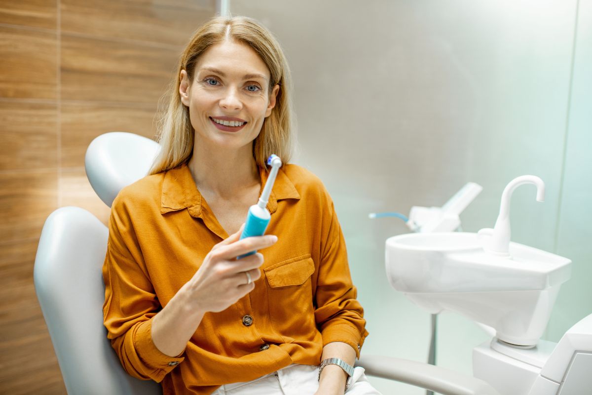 Woman in orange shirt holding toothbrush in modern dental clinic setting, smiling at camera.
