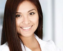 Professional headshot of woman with long dark hair wearing white blazer, smiling at camera indoors.